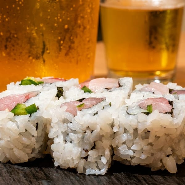 Six sushi rolls with seared tuna and green peppers on a dark plate, with two beers.