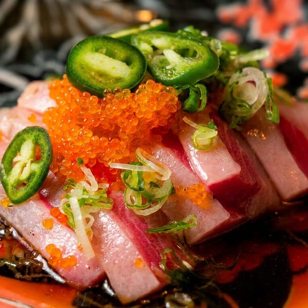 Sliced fish sashimi topped with orange roe, green jalapeños, and scallions on a decorative plate.
